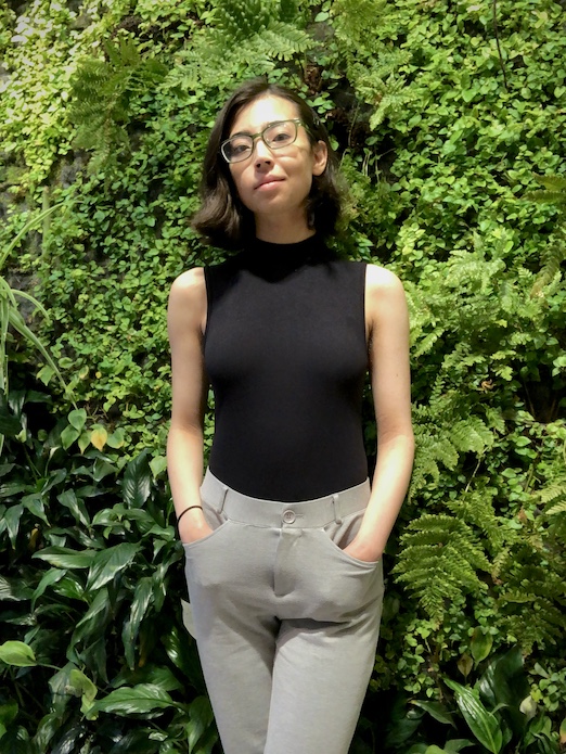 Anya, a Taiwanese-American guy with chin-length brown hair and green glasses, stands against a wall of lush green leaves. They are smiling and holding their hands in their pockets. They are wearing a sleeveless black turtleneck and gray dress pants.