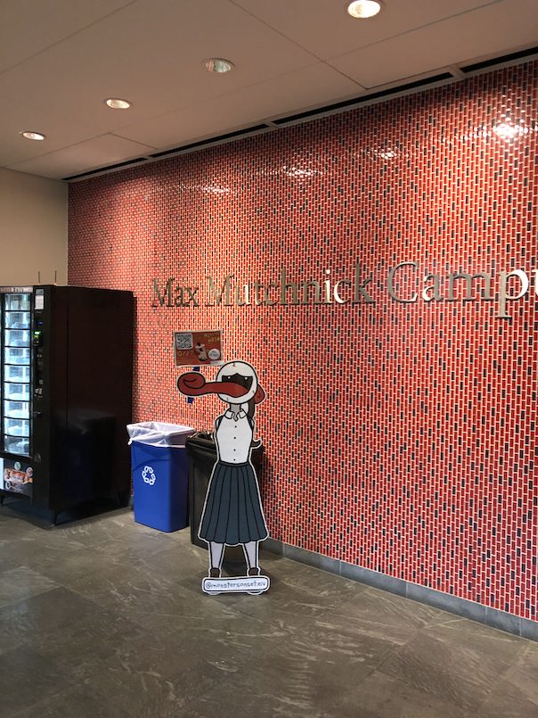 A photo of a cardboard cutout against a brick wall, next to a vending machine and a recycling bin. The cutout depicts a monster girl in a teal school uniform; she has an eyeball for a head and a long prehensile tongue, which she uses to hold a sign promoting a trivia event. Text by her feet reads '@monstersonset.eiv.'